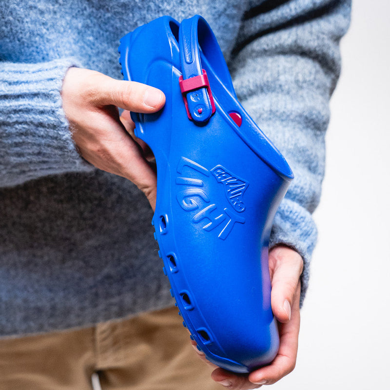A person in a gray knit sweater holds a bright blue clog-style shoe with a small red buckle detail against a plain light background.